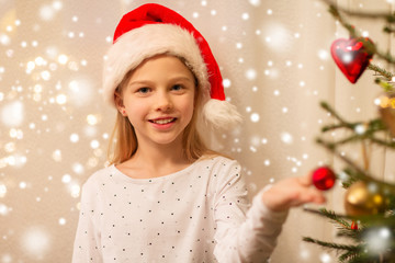 christmas, holidays and childhood concept - happy girl in santa hat decorating natural fir tree