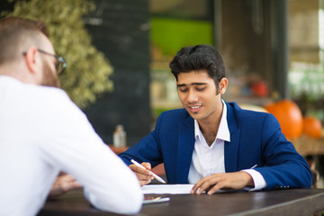 Smiling Indian businessman reading contract while meeting with partner in cafe. Content young business expert analyzing papers and editing it. Examining contract concept