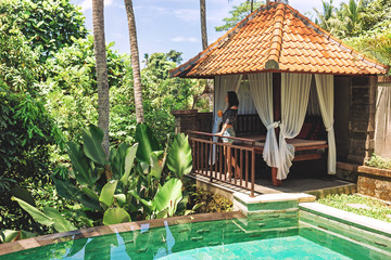 A young woman with a glass of juice in her hands is standing by the private pool against the backdrop of a romantic gazebo and tropical plants. A girl enjoys relaxing at a villa in Bali in Ubud.
