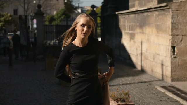 Pretty woman in black dress takes off the coat as walking down the street, outdoor shoot in old historical part of european city on great fall morning