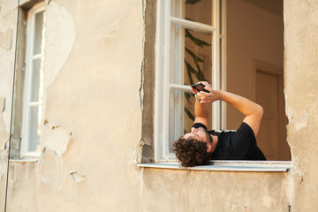 Cheerful young man texting and browsing the internet on his smart phone, and laying outside on the window taking a break