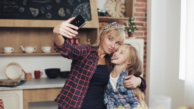 Smiling mom and daughter making selfie photo with smartphone camera at home in kitchen. Family, cook, and people concept