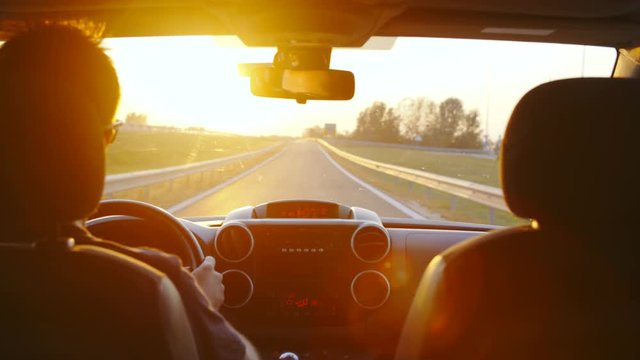Person drive the car towards the sunset in slow motion 4K. Static long shot from car back seat with dashboard and windshield in focus with view on the road. The person on left side driving.
