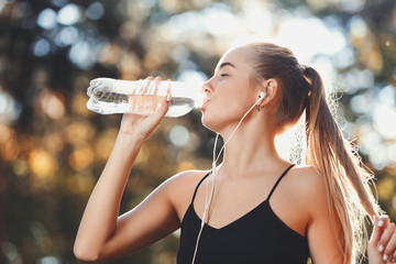 Portrait of young athletic long haired woman wearing sportish clothes listening music, touches her hair and drinking water in the sunny park, healthy lifestyle and people concept