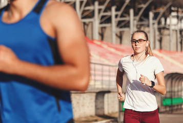 Sportish blonde long haired woman in white t-shirt and multicultural bearded man in blue shirt running a race at the football stadium outdoor, healthy lifestyle and people concept