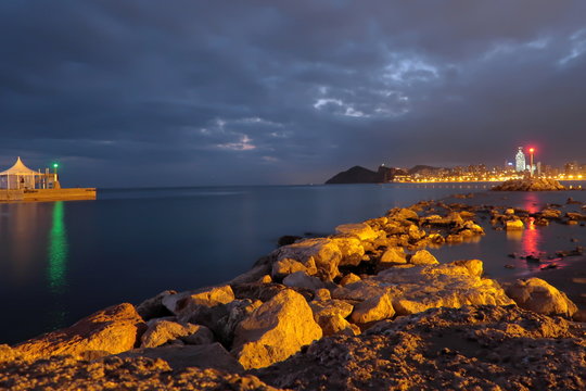 Night landscape on Poniente beach with rocks, lighthouse and buildings lights reflected in the water. Nightfall in Benidorm, Spain, with cloudy sky