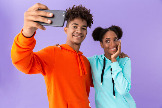 Photo of teenage african american couple wearing colorful sweatshirts taking selfie on smartphone, isolated over violet background