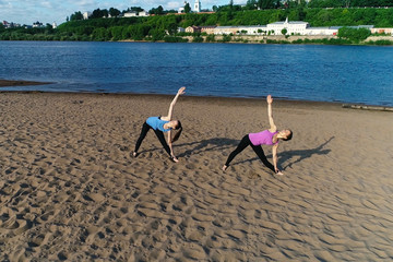 Two woman doing yoga on the sand beach by the river in the city. Beautiful city view in sunrise.