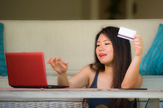 lifestyle portrait of young beautiful and happy Asian Chinese woman holding credit card banking and online shopping with laptop computer at home couch in internet business