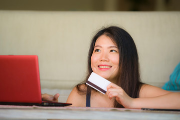 Fototapeta premium lifestyle portrait of young beautiful and happy Asian Korean woman holding credit card banking and online shopping with laptop computer at home couch in internet business