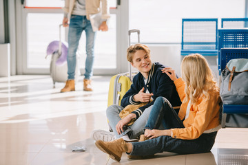 young people talking and smiling each other while sitting on floor and waiting for flight in airport