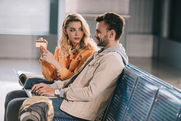 girl showing credit card to smiling man using laptop in airport