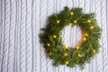 Fir wreath coiled with glowing garland with warm light on white knitted plaid.