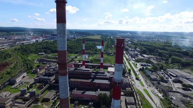 Cityscape with industrial building plant with three tall red and white industrial pipes over clear cloudless blue sky on sunny day. Footage. Top view near of white-red chimneys of plant. Concept of