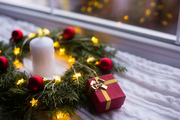 Gift box near festive decoration with red Christmas balls, white candle and warm garland lights.