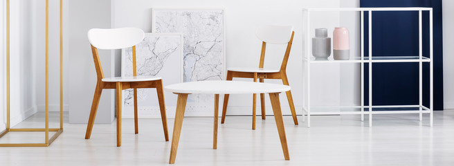 Panoramic view of two white wooden chairs and table, maps in frames and metal shelf in simple...