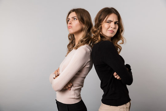 Image of two unhappy girls standing back to back with arms crossed and expressing resentment, isolated over gray background