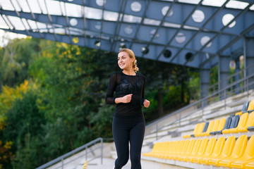 Smiling fit and sporty blonde woman in black sportswear jogging between the rows with yellow seats in the stadium. Healthy and sport concept. Morning training