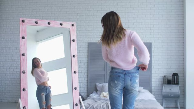 Beautiful adult woman in loose panrs admiring her body shape and appearance after losing weight while standing in front of big mirror in domestic room. Happy female showing weight loss success.