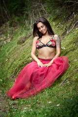 Beautiful brunette is sitting in the forest. She is wearing the flowered bra and red skirt. Glamour and fashion photography.