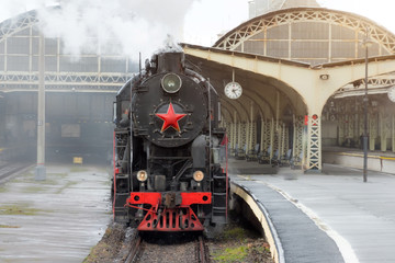 Retro steam train departs from the railway station with the clock on the platform.