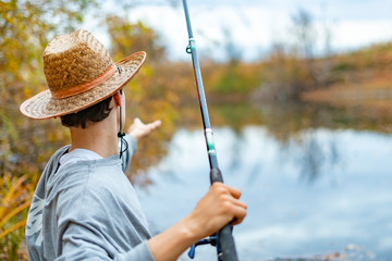 young man in hat sitting near lake and install set up and adjust fishing rod  f