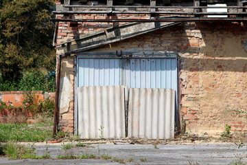Abandoned and in ruins red brick garage with metal doors blocked by large roof panels surrounded with rusted cracked gutter and wooden roof support with small brick wall and large trees in background