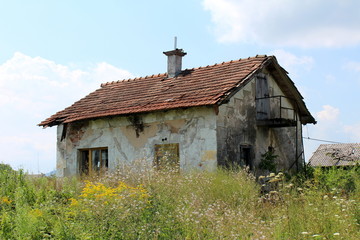 Fototapeta premium Burned small abandoned family house with completely dilapidated facade, destroyed doors and windows, broken and missing roof tiles surrounded with tall uncut grass mixed with flowers and cloudy blue s