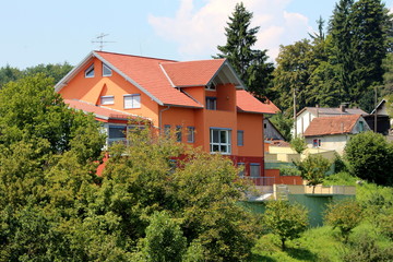 Modern suburban family house located on top of the hill surrounded with small older houses, tall trees and other forest vegetation on cloudy blue sky background