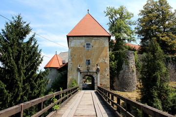 Obraz premium Wooden bridge entrance to old stone castle with dilapidated facade and completely new renovated roof surrounded with tall trees, overgrown vegetation and stone walls with cloudy blue sky background