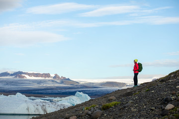 Obraz premium Young sporty girl with backpack admires beautiful view of the ice lake and mountains. Ice lagoon in Iceland