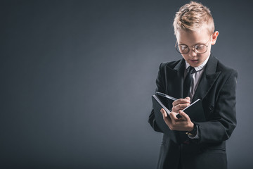 portrait of boy in businessman suit and eyeglasses making notes in notebook on grey backdrop