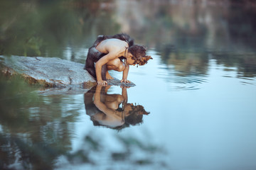 Caveman boy sitting on the rock and looking at him self in the water reflection in lake. Evolution survival concept. Creative art fantasy photo - obrazy, fototapety, plakaty