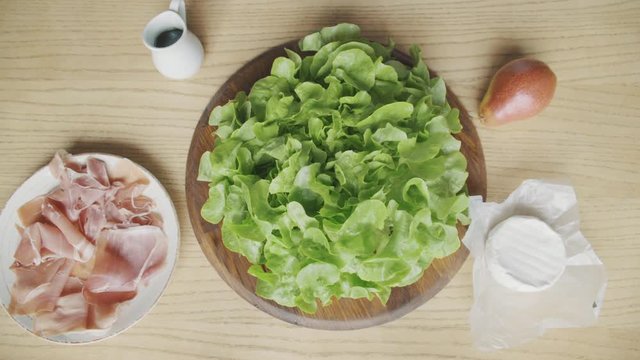 Male hands put salad ingredients on wooden table, top view, sauce, truffle oil, parma ham, jamon, pear, camembert, lettuce, salanova