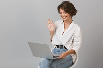 Happy young woman sitting on stool isolated over grey background holding laptop computer waving.