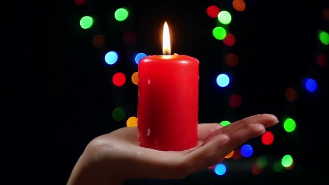 Close-up of female hand with a big red candle on bokeh background. Burning candle on a black background. Religion, spirituality and celebration.
