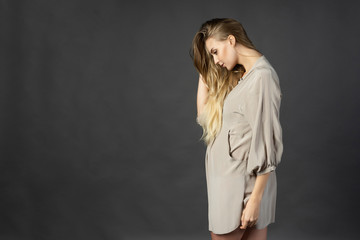 Fototapeta premium Beautiful young pregnant woman blonde, wearing a short light gray dress, stands sideways and touches her hair with her hand. Gray background. Maternity, lifestyle. Commercial design.