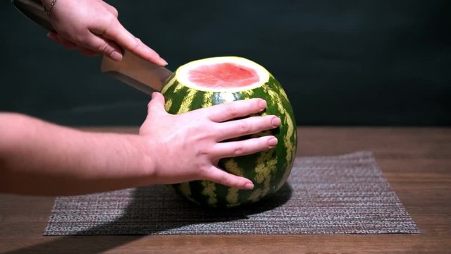 Watermelon is cut into slices with a knife. Wooden table. One red slice lays nearby.