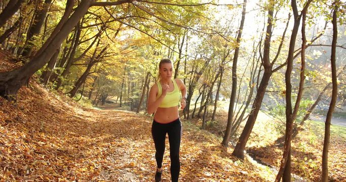 Female runner in the park. Woman looks straight in the camera while she runs along the path in the autumn park covered with fallen leaves