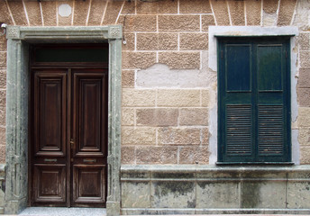 facade of an old spanish house with dark brown wooden door in a stone frame and black closed shutters with old crumbling textured wall tiles