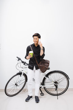 Photo of happy young asian man standing over white wall with bicycle. Looking camera drinking coffee.