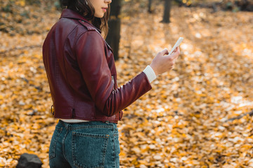 partial view of young woman in stylish leather jacket using smartphone in autumnal forest