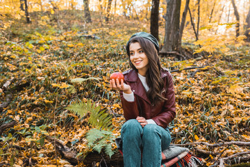 smiling stylish girl in leather jacket sitting on blanket and holding red apple in forest