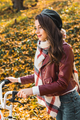 selective focus of cheerful woman in stylish leather jacket and beret carrying bicycle outdoors