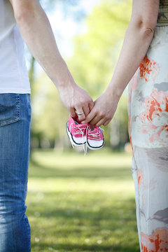 Parents Walk Together In Park, Holding Baby Shoes In Hands