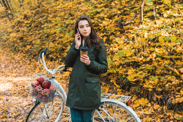 young woman with paper coffee cup talking on smartphone near bicycle in autumnal forest