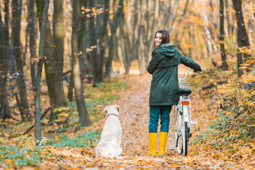 Fototapeta premium smiling woman with bicycle and her golden retriever sitting near on yellow leafy path in forest