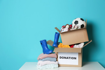 Donation boxes with toys, knitted clothes and shoes on table against color background. Space for...