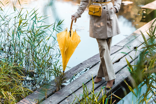cropped image of fashionable woman in trench coat standing with yellow umbrella near pond