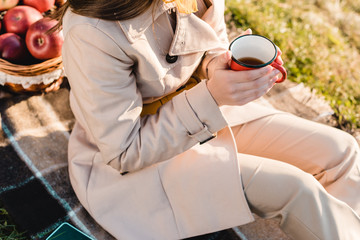 cropped image of fashionable young woman in trench coat holding cup of coffee while sitting on blanket outdoors